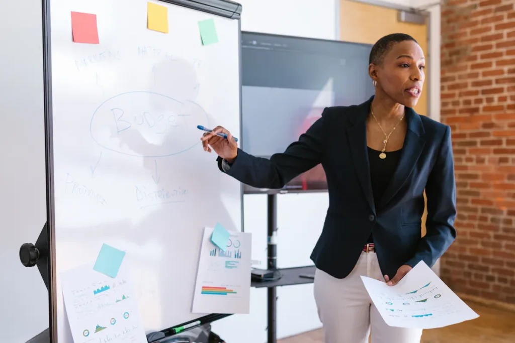 A woman in a blazer presents a budget plan on a whiteboard, highlighting the importance of financial planning in ESG training courses.